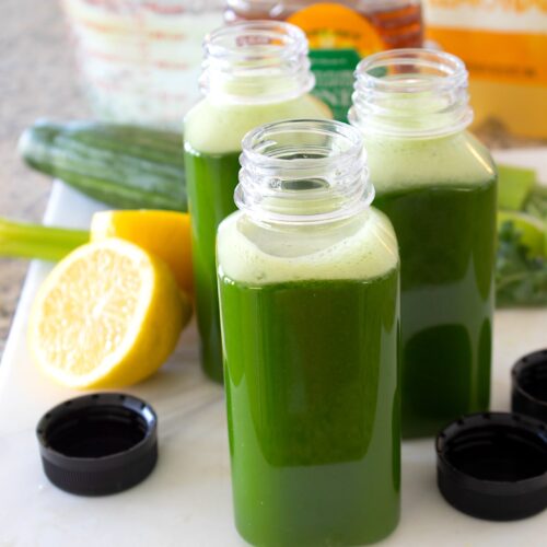 3 clear bottles filled with green juice and lemons and cucumbers in the background
